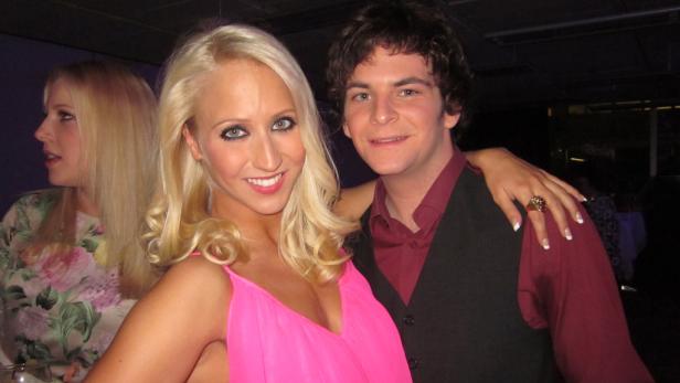 Eine blonde Frau im pinkfarbenen Kleid posiert mit einem Mann auf einer Party.