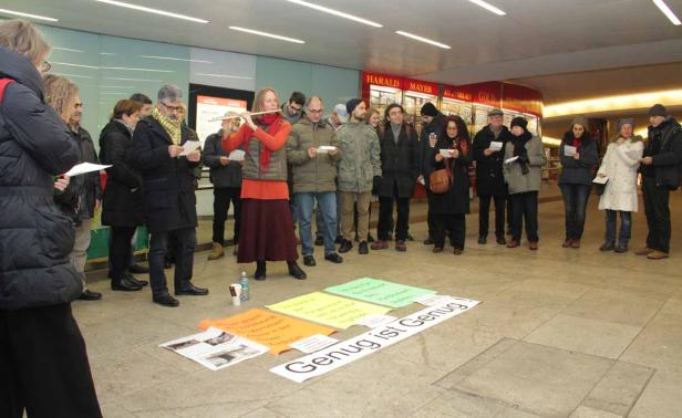 Eine Gruppe von Menschen steht in einer Halle, während eine Frau Flöte spielt und Schilder mit der Aufschrift „Genug ist Genug!“ auf dem Boden liegen.