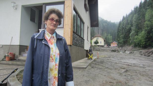 Eine ältere Frau steht vor einem Haus, dessen Hof von einer Überschwemmung verwüstet wurde.
