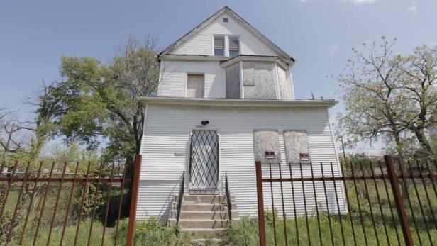 Ein verlassenes, weißes Haus mit vernagelten Fenstern und einem rostigen Zaun davor.