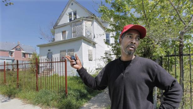 Ein Mann mit roter Kappe steht vor einem Haus mit vernagelten Fenstern.