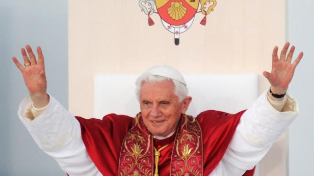 Papst Benedikt XVI. sitzt auf einem Thron und hebt die Hände.