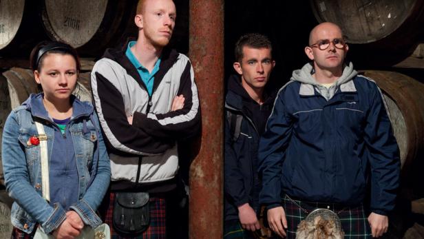 Vier Personen in Kilts stehen vor einer Wand aus Fässern.
