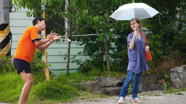Eine Frau mit Regenschirm und ein Mann in orangefarbenem T-Shirt stehen im Freien.