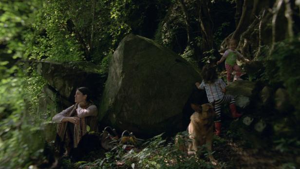 Eine Frau sitzt neben einem großen Felsen, während Kinder und ein Hund im Wald spielen.
