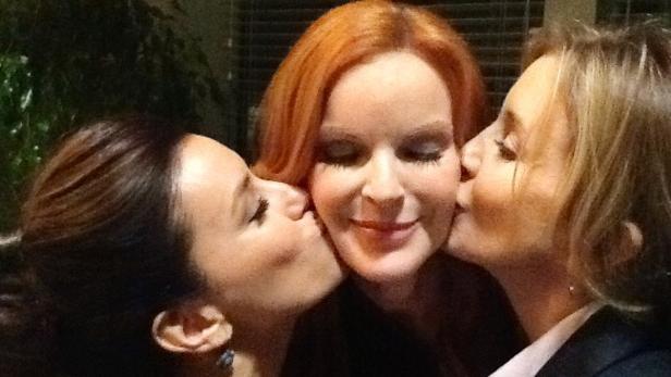 Zwei Frauen geben einer rothaarigen Frau gleichzeitig einen Kuss auf die Wange.