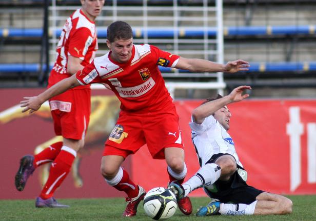 Ein Fußballspieler in Rot versucht, den Ball vor einem Gegenspieler zu schützen.