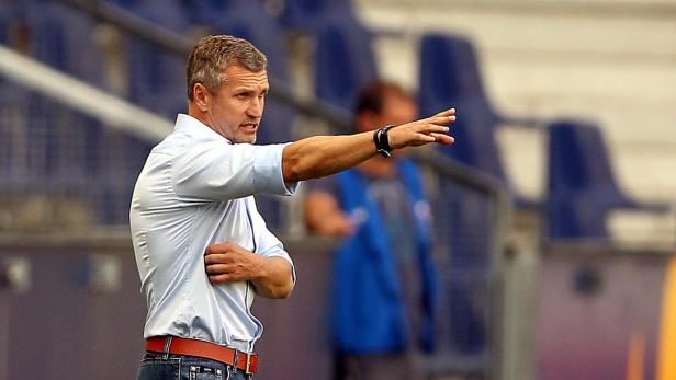 Ein Mann in Hemd und Jeans gestikuliert vor einer Tribüne.