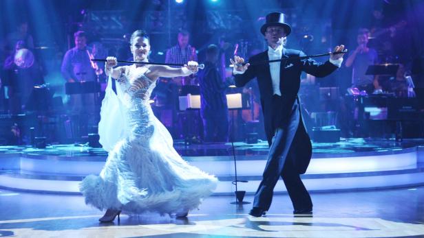 Ein Mann im Frack und eine Frau im Ballkleid tanzen auf einer Bühne mit Orchester im Hintergrund.