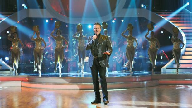 Ein Sänger steht mit Tänzerinnen und einem Orchester auf einer Bühne.