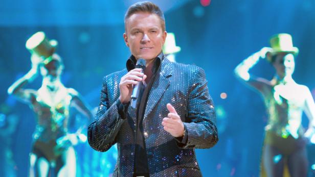 Ein Mann in einem glitzernden Sakko singt mit einem Mikrofon vor Tänzern mit Zylinderhüten.