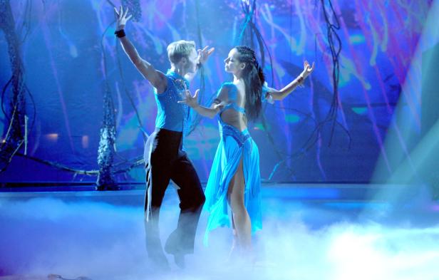 Ein Tanzpaar in blauen Kostümen auf einer Bühne mit Nebel und blauer Beleuchtung.