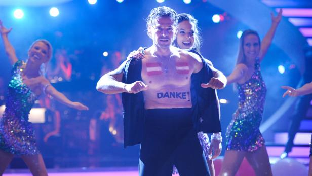 Ein Mann mit aufgemalter österreichischer Flagge und dem Wort „Danke!“ auf der Brust steht auf einer Bühne.