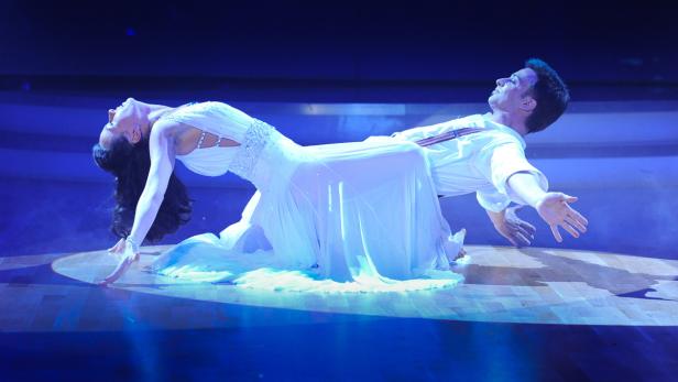Ein Tanzpaar in weißen Kostümen führt eine dramatische Pose auf einer Bühne auf.