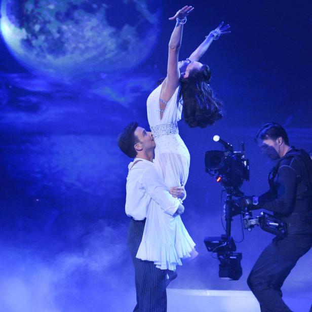 Ein Tänzer hebt seine Partnerin in einem weißen Kleid vor einem blauen Bühnenhintergrund hoch.