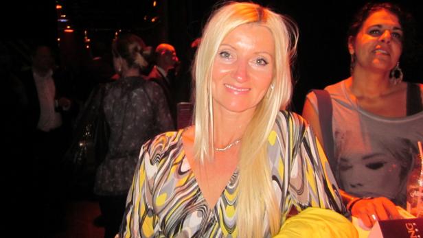 Eine blonde Frau lächelt in die Kamera, während sie in einem belebten Raum steht.