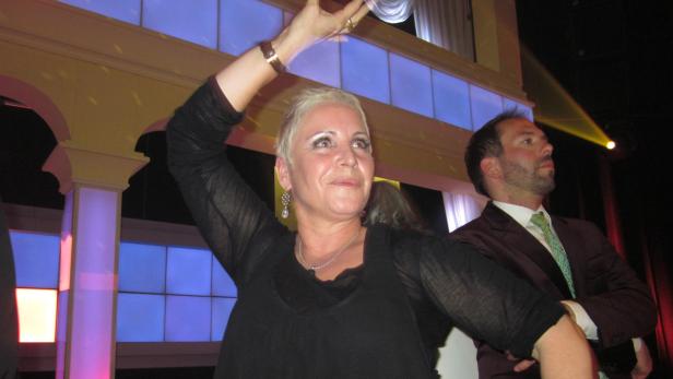 Eine Frau mit kurzem, blondem Haar tanzt auf einer Veranstaltung, während ein Mann im Anzug zusieht.