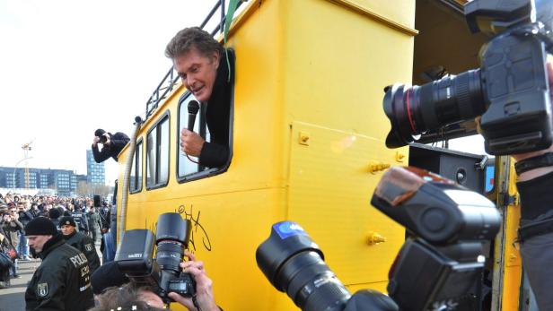 David Hasselhoff spricht aus einem gelben Bus zu einer Menschenmenge, umringt von Fotografen.