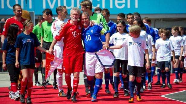 Franz Beckenbauer und ein weiterer Mann umarmen sich auf einem roten Teppich, umgeben von Kindern und anderen Personen.
