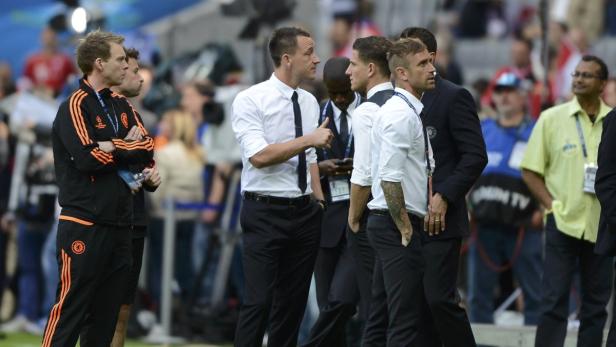Mehrere Männer, einige im Trainingsanzug, andere in Hemden und Anzügen, stehen auf einem Fußballfeld.