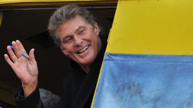 David Hasselhoff winkt aus einem gelb-blauen Fenster und hält einen Stift in der Hand.