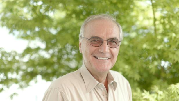 Ein lächelnder älterer Mann mit Brille vor grünem Hintergrund.