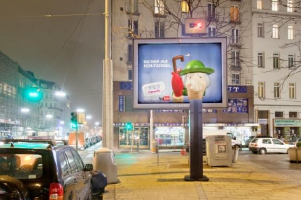 Pez/Rock+Partner/Gewista Ein beleuchteter Werbebildschirm auf einer Straße zeigt eine Comicfigur mit Regenschirm.