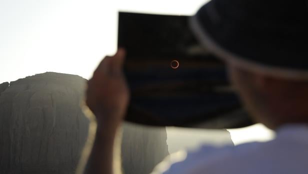 Eine Person betrachtet eine ringförmige Sonnenfinsternis durch ein dunkles Glas vor einer Felsformation.