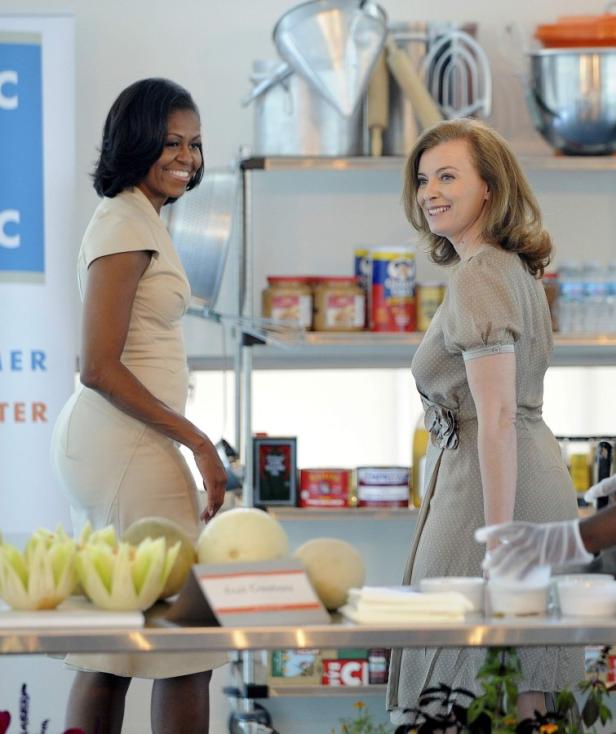 Michelle Obama und eine weitere Frau stehen in einer Küche mit Melonen und Konservenregalen.