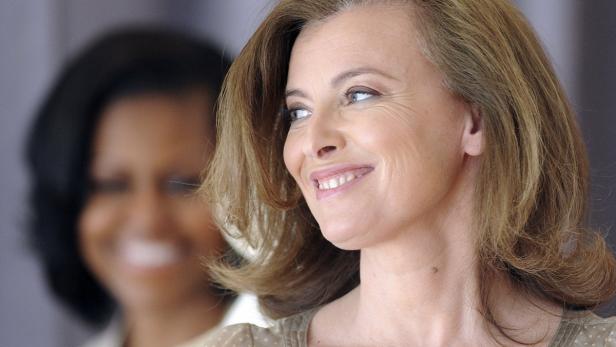 Valérie Trierweiler lächelt, während Michelle Obama im Hintergrund zu sehen ist.