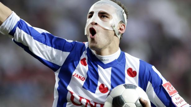 Ein Fußballspieler mit einer Gesichtsmaske jubelt mit einem Fußball in der Hand.