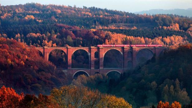 Ein roter Zug fährt über eine hohe Ziegelsteinbrücke, umgeben von herbstlichen Wäldern.