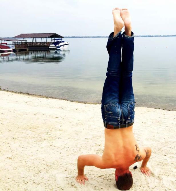 Ein Mann macht einen Kopfstand am Strand, im Hintergrund ein See mit einem Bootssteg.