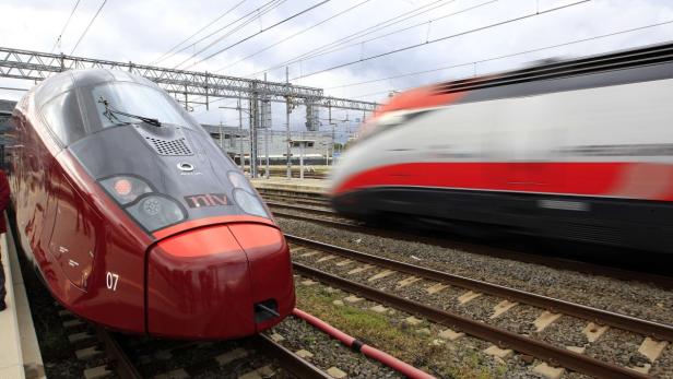 Ein roter Hochgeschwindigkeitszug der NTV steht auf einem Bahnhof, während ein anderer vorbeifährt.