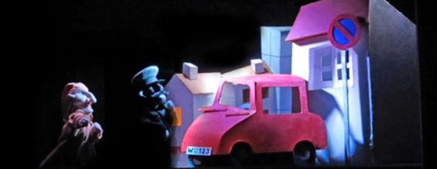 Eine Puppenspielszene mit einem roten Auto und einem Polizisten vor einem stilisierten Stadtbild.