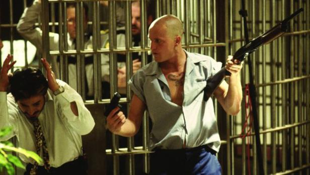 Ein Mann mit Tätowierung hält eine Schrotflinte und eine Pistole in einem Gefängnis.