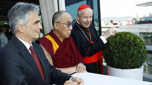 Der Dalai Lama und Kardinal Christoph Schönborn stehen mit einem Mann an einem Tisch auf einer Dachterrasse.
