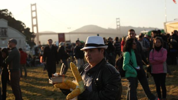 Ein Mann mit Hut hält Maiskolben und ein Dessert vor der Golden Gate Bridge.