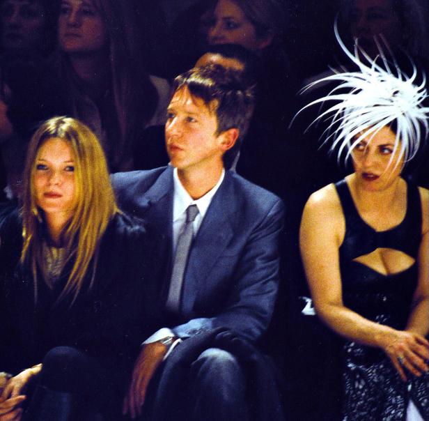 Kate Moss, ein Mann im Anzug und eine Frau mit extravagantem Hut sitzen in der ersten Reihe.