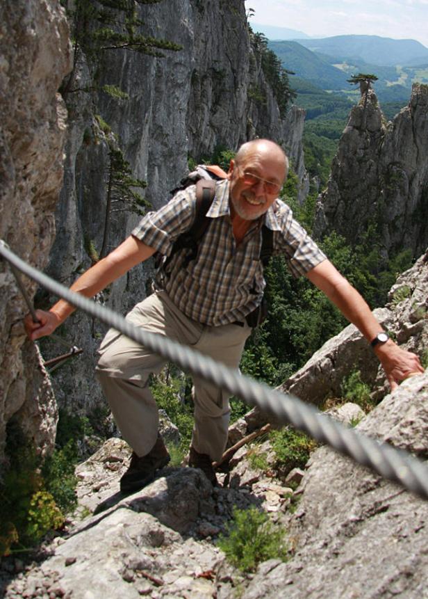 Ein älterer Mann klettert mit einem Seil gesichert einen felsigen Bergpfad hinauf.