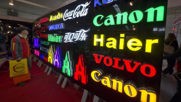 Ein Mann betrachtet eine Wand mit beleuchteten Logos verschiedener Marken wie Canon, Volvo und Coca-Cola.