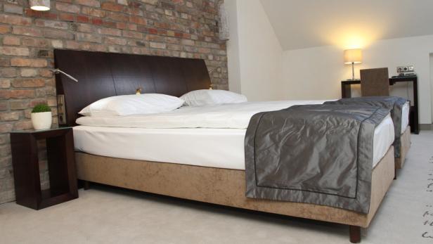 Ein Doppelbett mit grauer Tagesdecke in einem Hotelzimmer mit Ziegelwand.