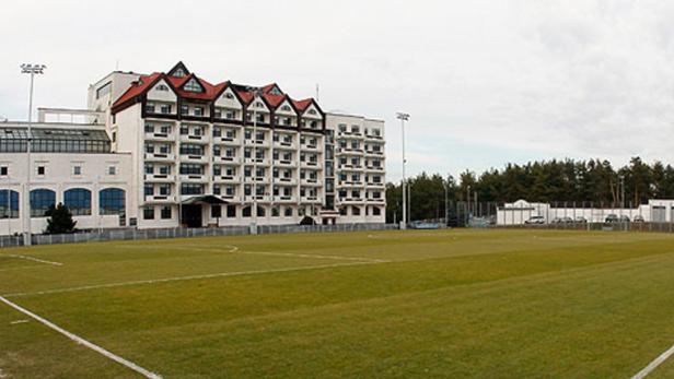 Ein großes, weißes Gebäude mit rotem Dach steht neben einem grünen Fußballfeld.