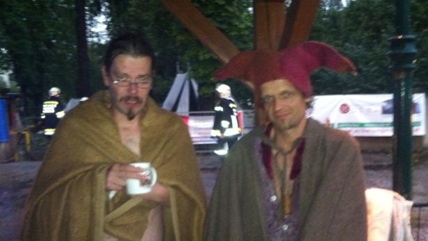 Zwei Männer, einer mit Narrenkappe, posieren; im Hintergrund ein Feuerwehrmann.