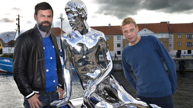 Zwei Männer stehen neben einer glänzenden Skulptur am Hafen.