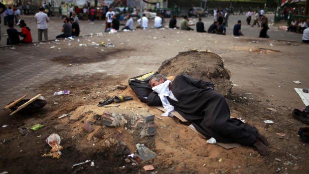 Ein Mann schläft mit einem Tuch um den Hals auf einem Sandhügel in der Öffentlichkeit.