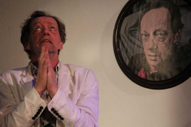 Ein Mann im weißen Sakko blickt mit gefalteten Händen nach oben, neben ihm ein Porträt.