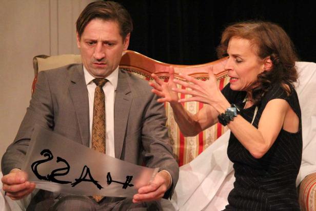 Eine Frau gestikuliert aufgeregt, während ein Mann mit einem beschrifteten Blatt Papier besorgt aussieht.