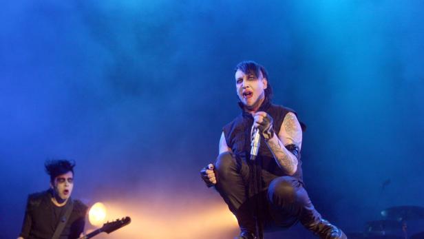 Marilyn Manson kniet mit Mikrofon auf der Bühne, während ein Gitarrist im Hintergrund spielt.