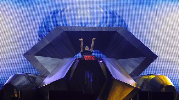 Ein DJ steht mit erhobenen Armen an einem futuristischen DJ-Pult vor einer blauen LED-Wand.